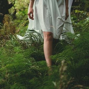 litha woman walking barefoot through ferns in a forest wearing a white dress